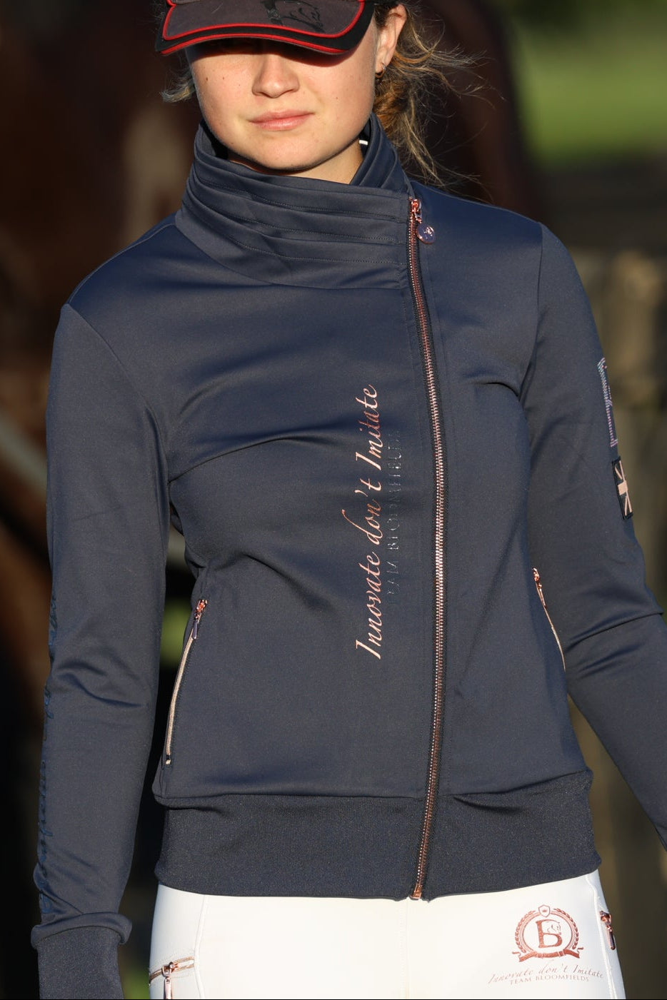 Woman wearing a navy blue jacket with a brand logo, standing in front of horses.